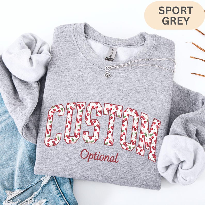 Pode incluir: Uma camisola de gola redonda cinzento desportivo com a palavra CUSTOM em letras grandes de bloco preenchidas com um padr&atilde;o de cerejas vermelhas e folhas verdes. Abaixo da palavra CUSTOM, a palavra Optional est&aacute; impressa numa fonte cursiva vermelha menor.