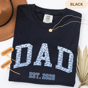 以下が含まれることがあります： イルカの模様で埋められた、ライトブルーの「DAD」という文字が入った黒いTシャツ。「EST. 2026」という文字が下にあります。金のネックレスと茶色の帽子が背景に見えます。シャツには「Comfort Colors」と書かれています。