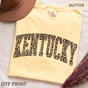 Peut inclure: T-shirt jaune clair avec le mot "KENTUCKY" en grandes lettres à imprimé léopard. Le t-shirt est étiqueté "BUTTER" et "XL - TG - EG". Les mots "DTF PRINT" sont en bas. Le t-shirt a une coupe décontractée.