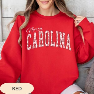 May include: A bright red crewneck sweatshirt featuring the text North CAROLINA printed in a large, arched font. The letters are filled with a white and red floral pattern. The garment has long sleeves and a ribbed neckline, cuffs, and hem.
