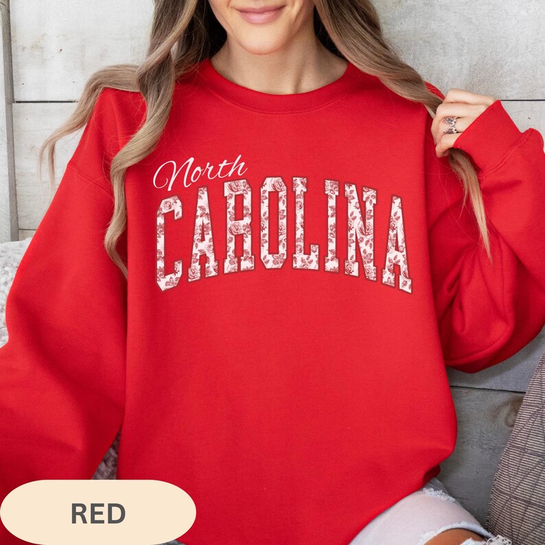 May include: A bright red crewneck sweatshirt featuring the text North CAROLINA printed in a large, arched font. The letters are filled with a white and red floral pattern. The garment has long sleeves and a ribbed neckline, cuffs, and hem.