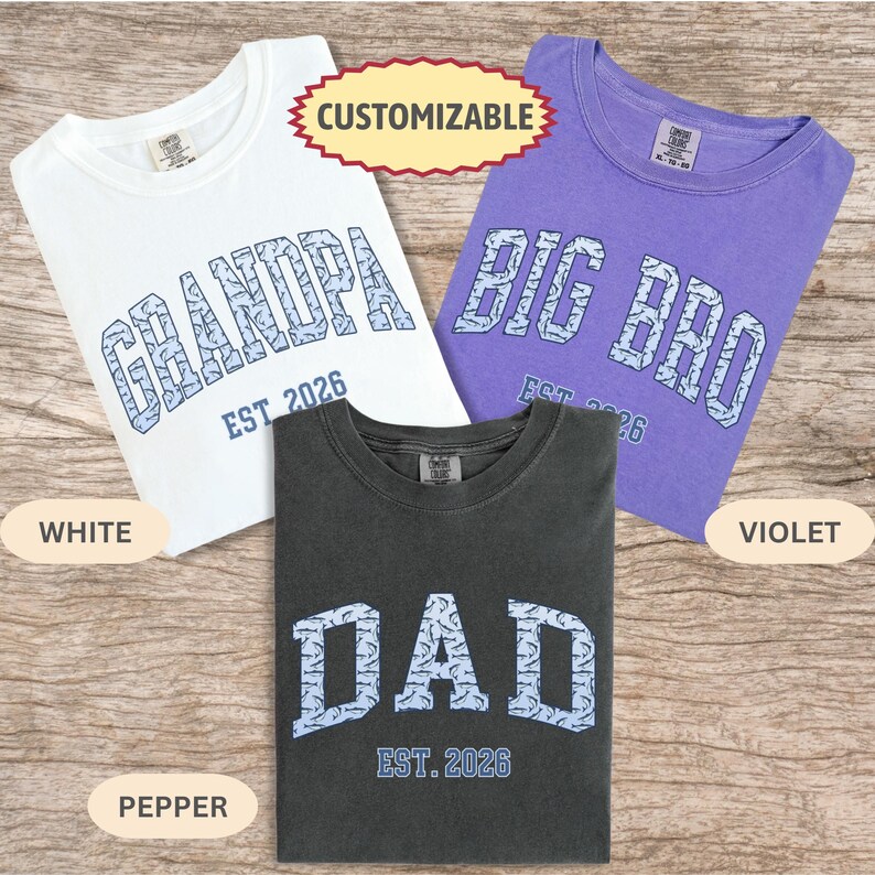 以下が含まれることがあります： 白、紫、ペッパーカラーの3枚のTシャツ。各シャツには、ライトブルーの色で異なるテキストデザインがあります。「GRANDPA」、「BIG BRO」、および「DAD」。テキスト「EST. 2026」は各単語の下に表示されます。「CUSTOMIZABLE」という単語も表示されます。