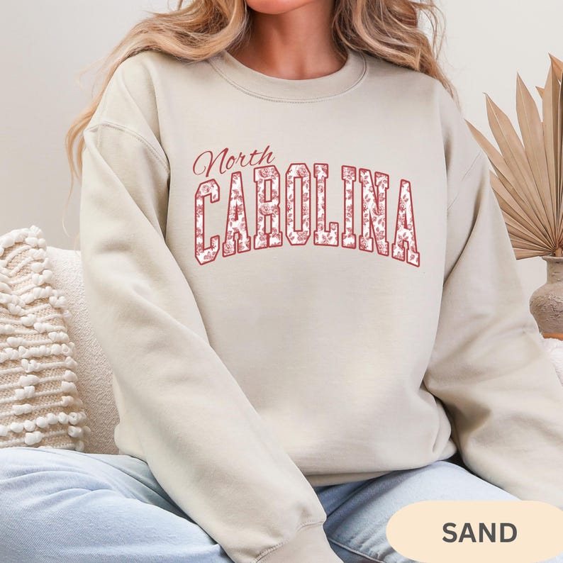 May include: A sand-colored crewneck sweatshirt featuring the text North CAROLINA in a collegiate font. The letters are outlined in red and filled with a detailed red floral pattern. The garment has long sleeves and a ribbed neckline and cuffs.