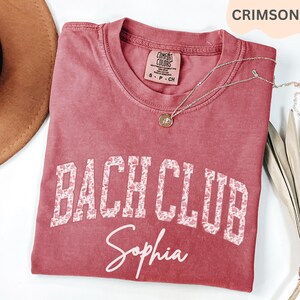 May include: Crimson-colored t-shirt with the words "BACH CLUB" in a floral design and the name "Sophia" in script. The shirt has a crew neck and short sleeves. A gold necklace with a pendant is draped on the shirt.