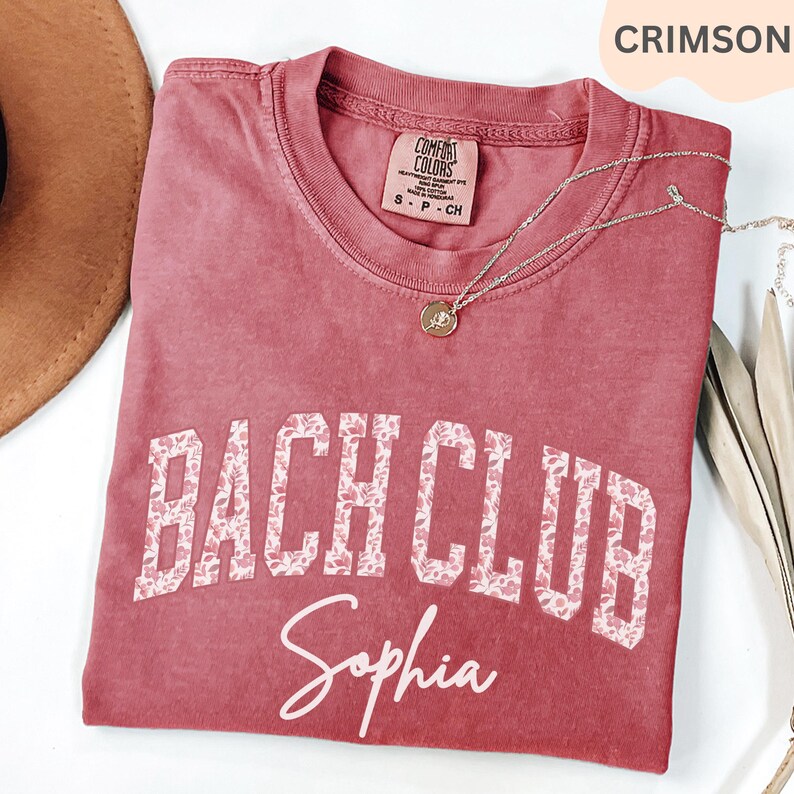 May include: Crimson-colored t-shirt with the words "BACH CLUB" in a floral design and the name "Sophia" in script. The shirt has a crew neck and short sleeves. A gold necklace with a pendant is draped on the shirt.