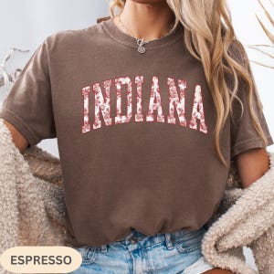 以下が含まれることがあります： 「INDIANA」の文字がフローラルデザインで描かれたエスプレッソカラーのTシャツ。テキストは、赤いアウトラインと白いフローラルパターンを持つカレッジスタイルのフォントです。シャツは無地で、半袖です。