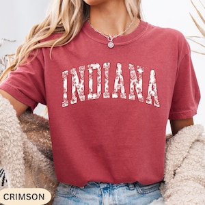 以下が含まれることがあります： 「INDIANA」の文字が白い花柄でプリントされたクリムゾンカラーのTシャツ。Tシャツは半袖でクルーネックです。「CRIMSON」の文字が左下の白い長方形にあります。