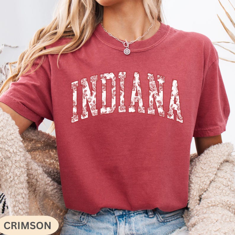 以下が含まれることがあります： 「INDIANA」の文字が白い花柄でプリントされたクリムゾンカラーのTシャツ。Tシャツは半袖でクルーネックです。「CRIMSON」の文字が左下の白い長方形にあります。