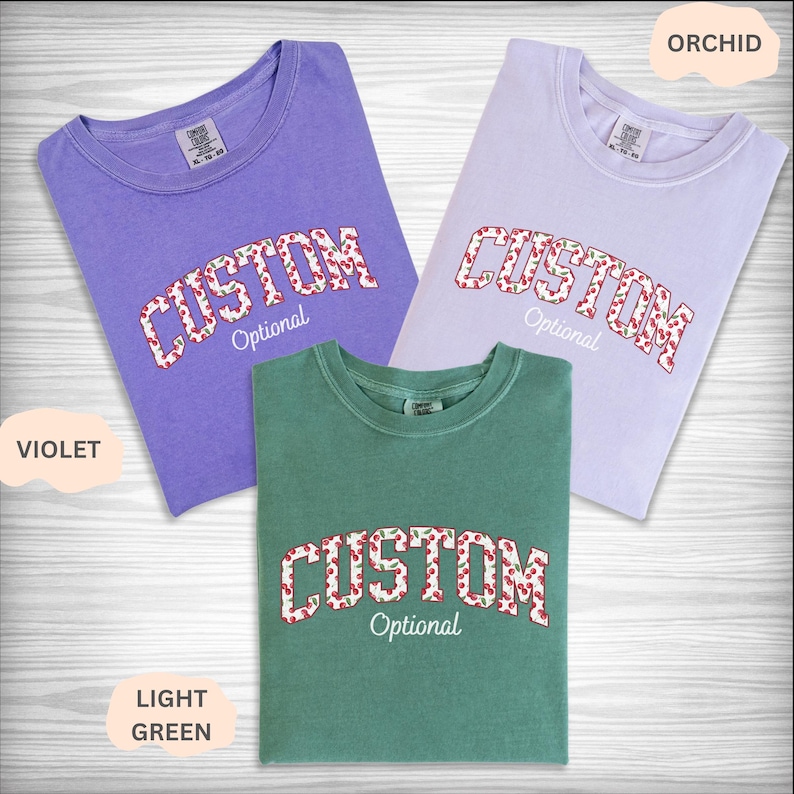Pu&ograve; includere: Tre magliette a maniche corte con girocollo in viola, orchidea e verde chiaro. Ogni maglietta presenta la parola CUSTOM in un carattere con motivo a ciliegie e contorno bianco, con la parola Optional in corsivo bianco sotto il testo.