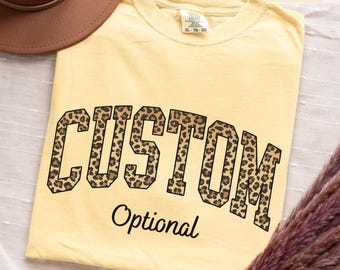 May include: A butter yellow t-shirt featuring the word CUSTOM in a large, block-style font with a brown and black leopard print pattern, followed by the word Optional in a smaller, black cursive script font, with a size label reading XL-TG-EG.