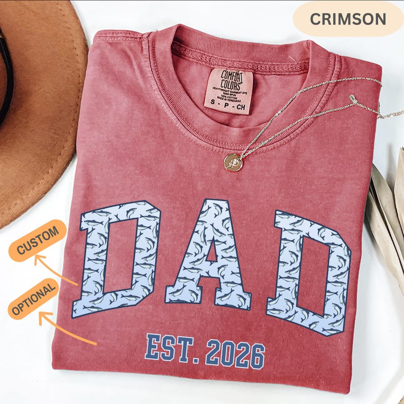 以下が含まれることがあります： 「DAD」の文字がサメの模様で埋められた、明るい青色のブロック体で書かれたクリムゾン色のTシャツ。「EST. 2026」の文字が「DAD」の下にあります。ペンダント付きのゴールドネックレスがシャツの上にあります。