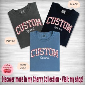 Pu&ograve; includere: Tre magliette a girocollo nei colori pepper, nero e blue jean. Ogni maglietta presenta la parola CUSTOM in un carattere con motivo a ciliegie, con la parola Optional scritta sotto in corsivo bianco. Le magliette sono disposte su una superficie in legno chiaro.