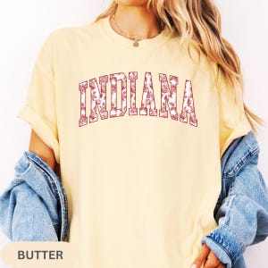 以下が含まれることがあります： 薄い黄色のTシャツ。前面に、花柄の模様が入ったマルーン色のフォントで「INDIANA」と書かれています。クルーネックと半袖のデザインです。「BUTTER」という文字が画像の下部にあります。