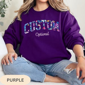 May include: A deep purple crewneck sweatshirt featuring the word CUSTOM in large, block letters filled with a colorful floral pattern. Below the word CUSTOM, the word Optional is printed in a white, cursive script font on the chest area.