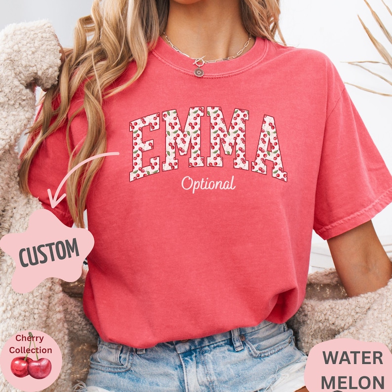 Pu&ograve; includere: Una t-shirt a maniche corte color corallo con il nome EMMA in grandi lettere stampatello riempite con un motivo a ciliegie rosse. Sotto il nome, la parola Optional &egrave; stampata in corsivo bianco. La maglietta &egrave; indossata con jeans blu.