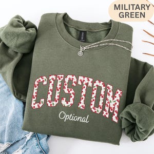 Pode incluir: Uma camisola de gola redonda verde militar com a palavra CUSTOM em letras de bloco preenchidas com um padr&atilde;o de cerejas vermelhas e a palavra Optional em letra cursiva branca por baixo. Um colar de prata repousa na gola e uma etiqueta diz GILDAN.