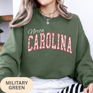 May include: A military green crewneck sweatshirt featuring the text North CAROLINA in a bold, arched font. The letters are filled with a white and red floral pattern and outlined in red. The sweatshirt has long sleeves and a ribbed neckline.