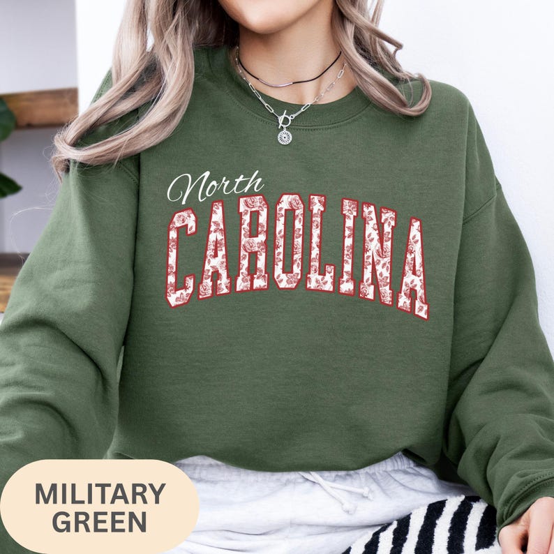 May include: A military green crewneck sweatshirt featuring the text North CAROLINA in a bold, arched font. The letters are filled with a white and red floral pattern and outlined in red. The sweatshirt has long sleeves and a ribbed neckline.
