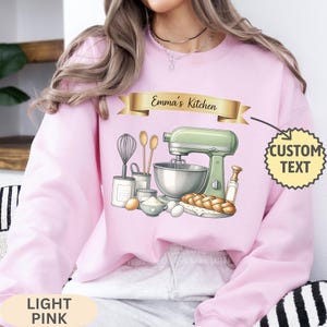 May include: A light pink sweatshirt featuring a graphic of a light green stand mixer, a whisk, wooden spoons, eggs, flour, and a braided loaf of bread. A gold banner above the graphic displays the text Emma's Kitchen, with a starburst label indicating custom text.