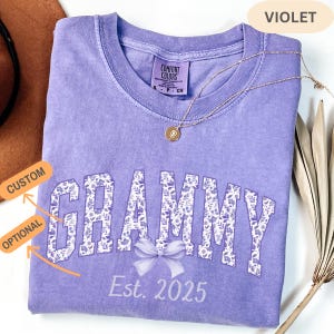May include: A violet Comfort Colors t-shirt with the word "GRAMMY" in a floral design, a bow, and the text "Est. 2025". A gold necklace with a pendant is draped across the shirt.