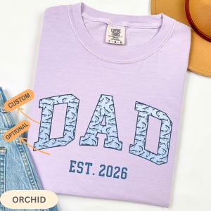以下が含まれることがあります： 紫色のTシャツ。青いフォントで「DAD」の文字が魚の模様で埋められています。「EST. 2026」の文字が「DAD」の下にあります。シャツにはComfort Colorsのタグが付いています。