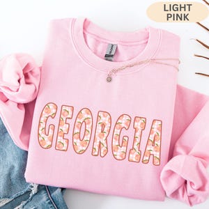 May include: A light pink crewneck sweatshirt featuring the word GEORGIA in large block letters. The letters have a white and pink gingham background pattern overlaid with small peach illustrations. A silver chain necklace rests on the collar.