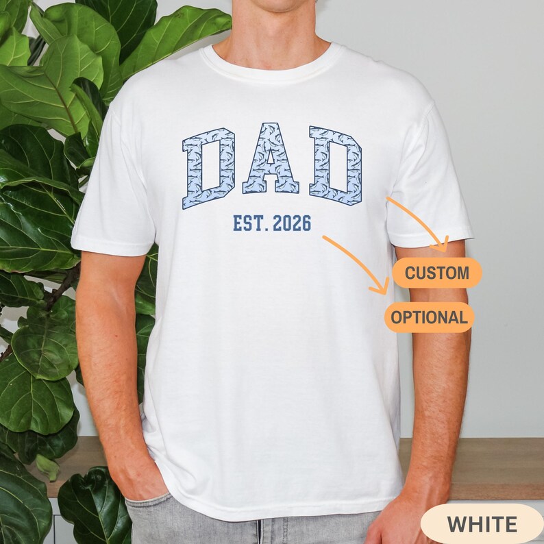 以下が含まれることがあります： 青いテクスチャード加工の「DAD」という文字と「EST. 2026」というテキストがプリントされた白いTシャツ。Tシャツは半袖でクルーネックです。デザインは胸の中央に配置されています。