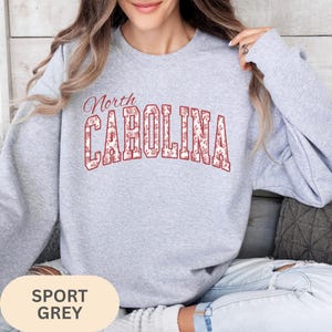 May include: A light grey crewneck sweatshirt featuring the text North CAROLINA in a bold, arched font. The letters are filled with a detailed red and white floral pattern. The sweatshirt has long sleeves and a ribbed collar, cuffs, and hem.