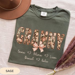 May include: Sage green t-shirt with the word "GRANNY" in a floral design. Below the word are the names Emma, Olivia, Daniel, Liam, and Katie, with hearts. A light brown hat is in the background.