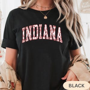 以下が含まれることがあります： 白い文字で「INDIANA」と赤い花柄がプリントされた黒いTシャツ。Tシャツは半袖でクルーネックです。「BLACK」の文字が右下に表示されています。