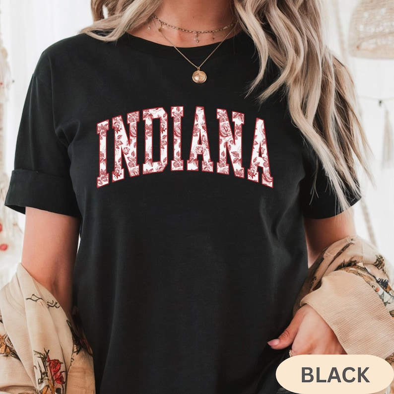 以下が含まれることがあります： 白い文字で「INDIANA」と赤い花柄がプリントされた黒いTシャツ。Tシャツは半袖でクルーネックです。「BLACK」の文字が右下に表示されています。