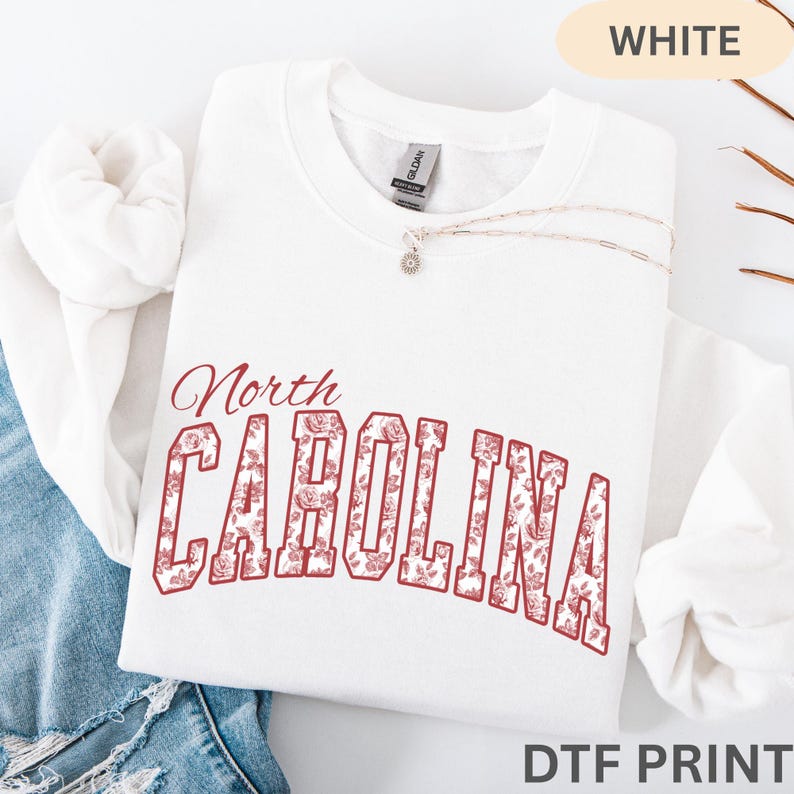 May include: A white crewneck sweatshirt featuring the text North CAROLINA in a red floral pattern. The word North is in a script font above the block-lettered CAROLINA, which is filled with a detailed rose print. A gold chain necklace rests on the collar.