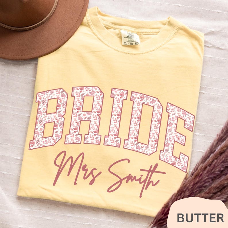 May include: Butter yellow t-shirt with the word "BRIDE" in large, floral-patterned letters. Below, the name "Mrs Smith" is written in a cursive font. The shirt is a size XL-TG-EG.