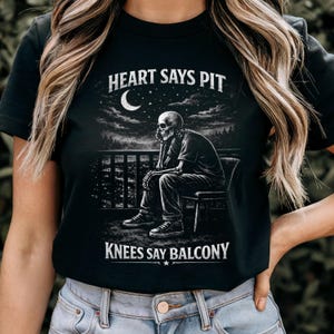 Pu&ograve; includere: T-shirt nera con grafica di uno scheletro seduto su una sedia, con la scritta "HEART SAYS PIT" sopra e "KNEES SAY BALCONY" sotto. Il design include un cielo notturno con una luna crescente e stelle.