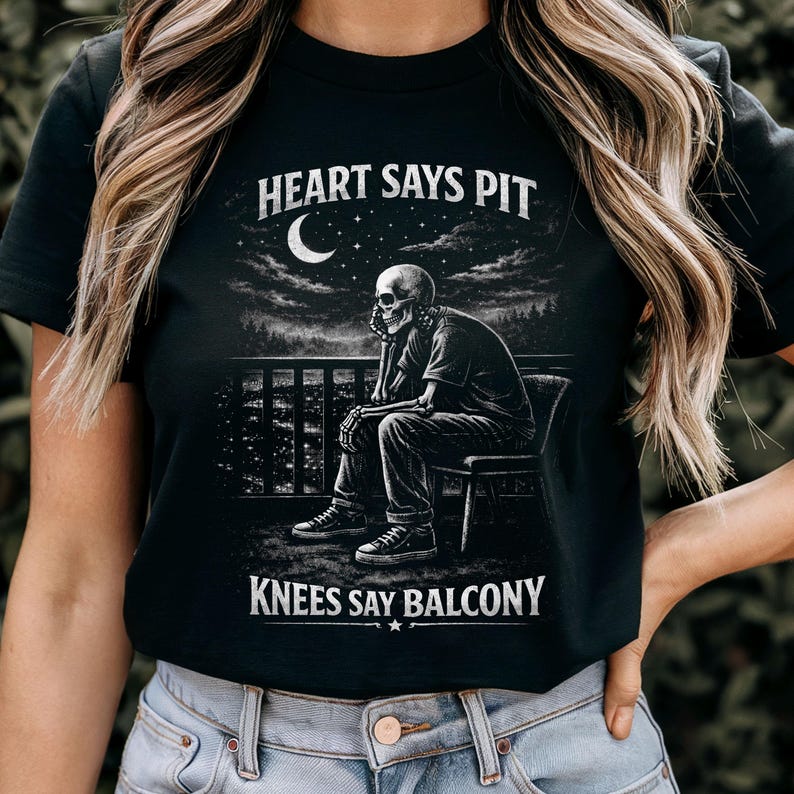 Pu&ograve; includere: T-shirt nera con grafica di uno scheletro seduto su una sedia, con la scritta "HEART SAYS PIT" sopra e "KNEES SAY BALCONY" sotto. Il design include un cielo notturno con una luna crescente e stelle.