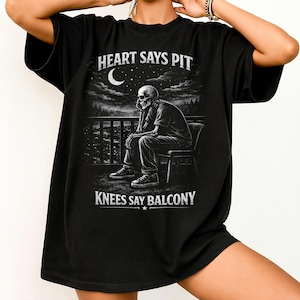 Pu&ograve; includere: T-shirt nera con grafica in scala di grigi di uno scheletro seduto su un balcone, con luna e paesaggio urbano sullo sfondo. Il testo "HEART SAYS PIT" e "KNEES SAY BALCONY" &egrave; stampato in bianco.