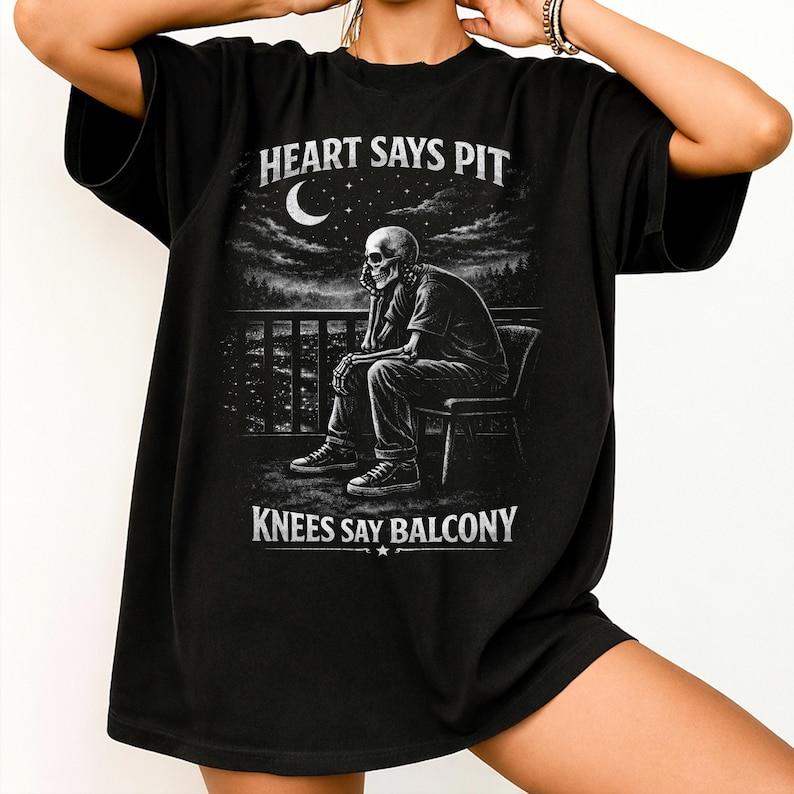 Pu&ograve; includere: T-shirt nera con grafica in scala di grigi di uno scheletro seduto su un balcone, con luna e paesaggio urbano sullo sfondo. Il testo "HEART SAYS PIT" e "KNEES SAY BALCONY" &egrave; stampato in bianco.
