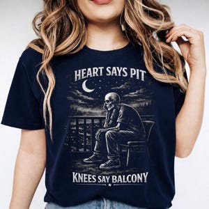 Pu&ograve; includere: T-shirt blu navy con grafica di uno scheletro seduto su un balcone sotto una luna a mezzaluna. Il testo recita "HEART SAYS PIT" e "KNEES SAY BALCONY". Il design &egrave; in bianco e grigio.