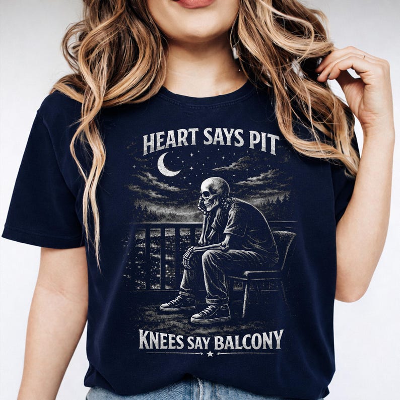 Pu&ograve; includere: T-shirt blu navy con grafica di uno scheletro seduto su un balcone sotto una luna a mezzaluna. Il testo recita "HEART SAYS PIT" e "KNEES SAY BALCONY". Il design &egrave; in bianco e grigio.