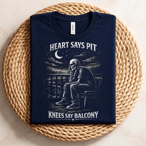 Pu&ograve; includere: T-shirt blu navy con grafica in bianco e nero di uno scheletro seduto su un balcone, con la scritta "HEART SAYS PIT" e "KNEES SAY BALCONY". Il design &egrave; ambientato su uno sfondo di cielo notturno.