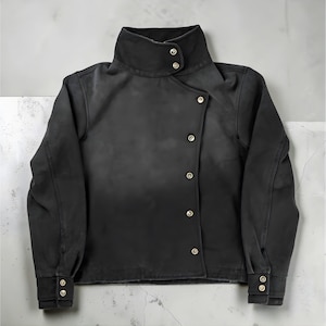 Op de afbeelding: Zwarte jas met een hoge kraag en goudkleurige knopen. De jas heeft een asymmetrische knoopsluiting en manchetten met knopen. De stof lijkt enigszins versleten of vervaagd.