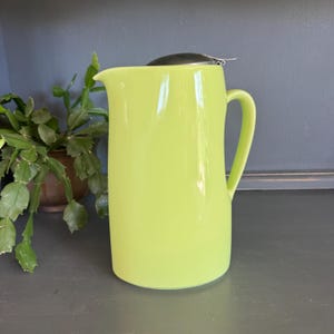 May include: A bright lime green pitcher with a curved handle and a spout. A stainless steel tea infuser sits on top. The pitcher is on a dark gray surface, with a potted plant in the background.