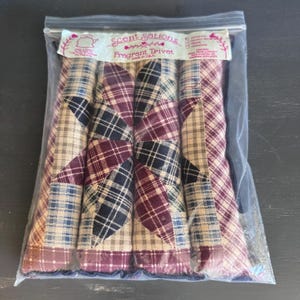 May include: A package of fragrant trivets with a patchwork design. The trivets feature a star pattern in shades of burgundy, navy, tan, and blue plaid fabric. The package is labeled "Scent Sations Fragrant Trivet" and "made in USA."