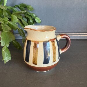 May include: A small, ceramic pitcher with a cream-colored body and vertical stripes in navy blue and brown. The rim and base are brown, and the handle is a darker brown. The pitcher has a spout for easy pouring and is likely used for serving liquids.