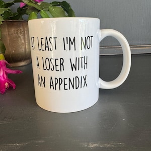 May include: White ceramic mug with a curved handle. The mug features black text that reads "AT LEAST I'M NOT A LOSER WITH AN APPENDIX". The mug is on a dark gray surface, with a plant in the background.