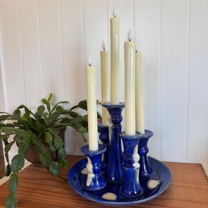 May include: A blue ceramic candelabra with six cream-colored taper candles. The candelabra has a round base and five candle holders. The candle holders are blue with white accents. The candles are approximately 10 inches tall.