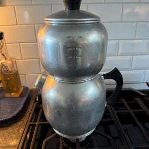 May include: A vintage aluminum coffee percolator with a black handle and lid knob. The percolator has measurement markings for 6, 9, and 12 cups. It sits on a black gas stovetop.