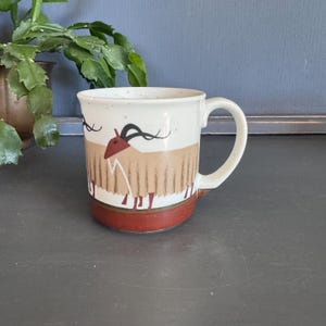 May include: Cream-colored ceramic mug with a brown and red design. The mug features a stylized illustration of a goat with black horns and a red head. The bottom of the mug is a dark red color.