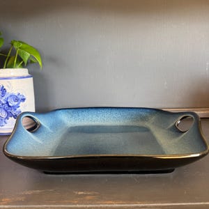 May include: A rectangular ceramic serving dish with a gradient blue interior and black exterior. The dish has two circular handles and a gold trim. The dish is on a dark surface.