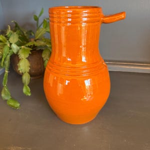 May include: A glossy orange ceramic pitcher with a handle. The pitcher has a bulbous body and a narrow neck, with horizontal ridges near the top. The handle extends horizontally from the side. A houseplant is in the background.